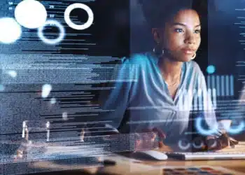 Mulher trabalhando em um computador com gráficos digitais e código em segundo plano, representando análise de dados.