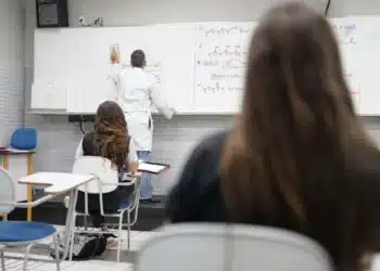 Estudantes assistem a aula de química em sala de aula, enquanto o professor escreve na lousa.