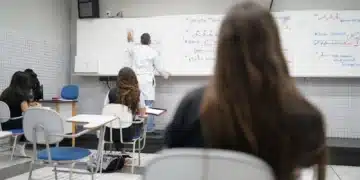 Estudantes assistem a aula de química em sala de aula, enquanto o professor escreve na lousa.