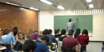 Aula em sala de aula com alunos e professor escrevendo no quadro verde.