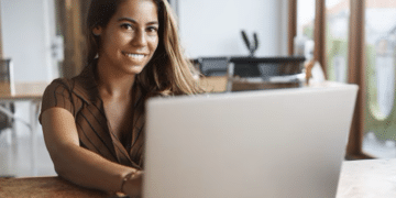 Mulher sorrindo enquanto trabalha em um laptop em ambiente moderno e iluminado.