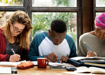 Três pessoas estudando em uma mesa, com livros, cadernos e canetas, em ambiente com luz natural.