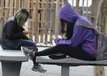 Estudantes sentadas em banco ao ar livre usando celulares.