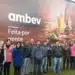 Funcionários da Ambev posando sorridentes em frente a um grande painel institucional com a frase "Feita por gente apaixonada."