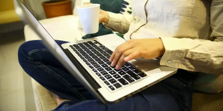 Pessoa trabalhando de casa, usando um laptop e segurando uma xícara de café.