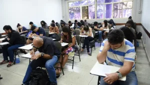 Grupo de estudantes em uma sala de aula realizando uma prova em carteiras individuais.