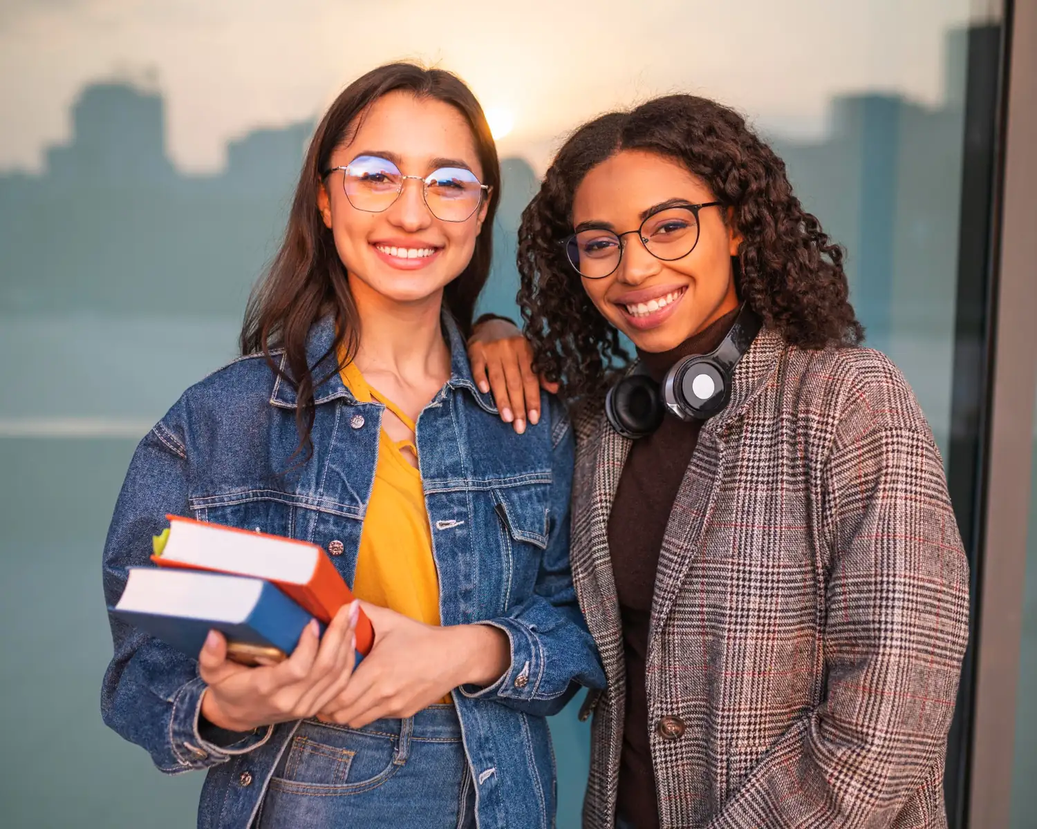 Duas estudantes sorrindo, uma segurando livros e outra com fones de ouvido no pescoço.