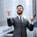 Homem de terno comemorando com expressão de sucesso em frente a um prédio moderno.