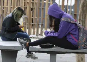 Duas jovens sentadas em um banco de concreto ao ar livre, concentradas no celular, vestindo roupas casuais.