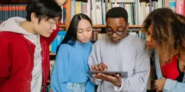 Grupo de estudantes analisando um tablet em ambiente acadêmico com prateleiras de livros ao fundo.
