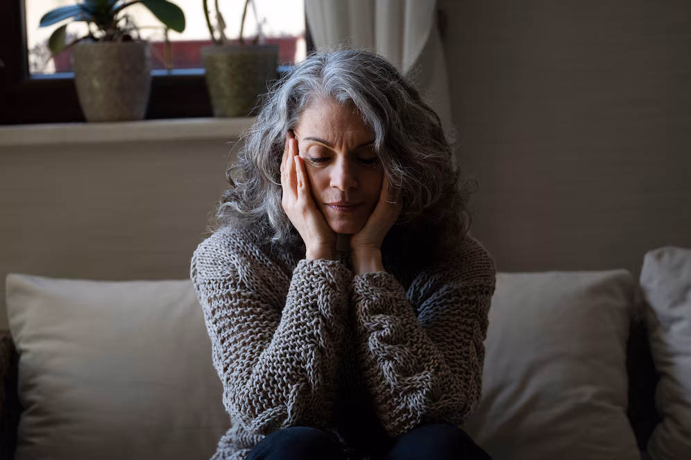 Mulher mais velha com expressão triste, sentada no sofá, aparentando estar sobrecarregada emocionalmente.