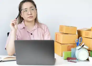 Pessoa pensando enquanto trabalha em frente a um laptop com caixas empilhadas ao fundo.