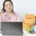Pessoa pensando enquanto trabalha em frente a um laptop com caixas empilhadas ao fundo.