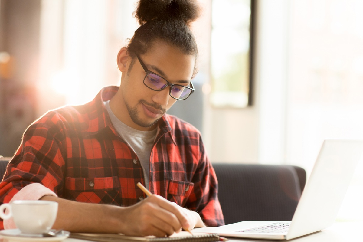 Homem jovem estudando com laptop e caderno, com uma xícara de café na mesa, em ambiente claro e arejado.