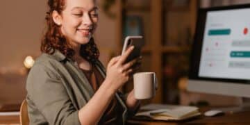 Mulher feliz segurando celular e caneca, trabalhando confortavelmente de casa com computador ao fundo.