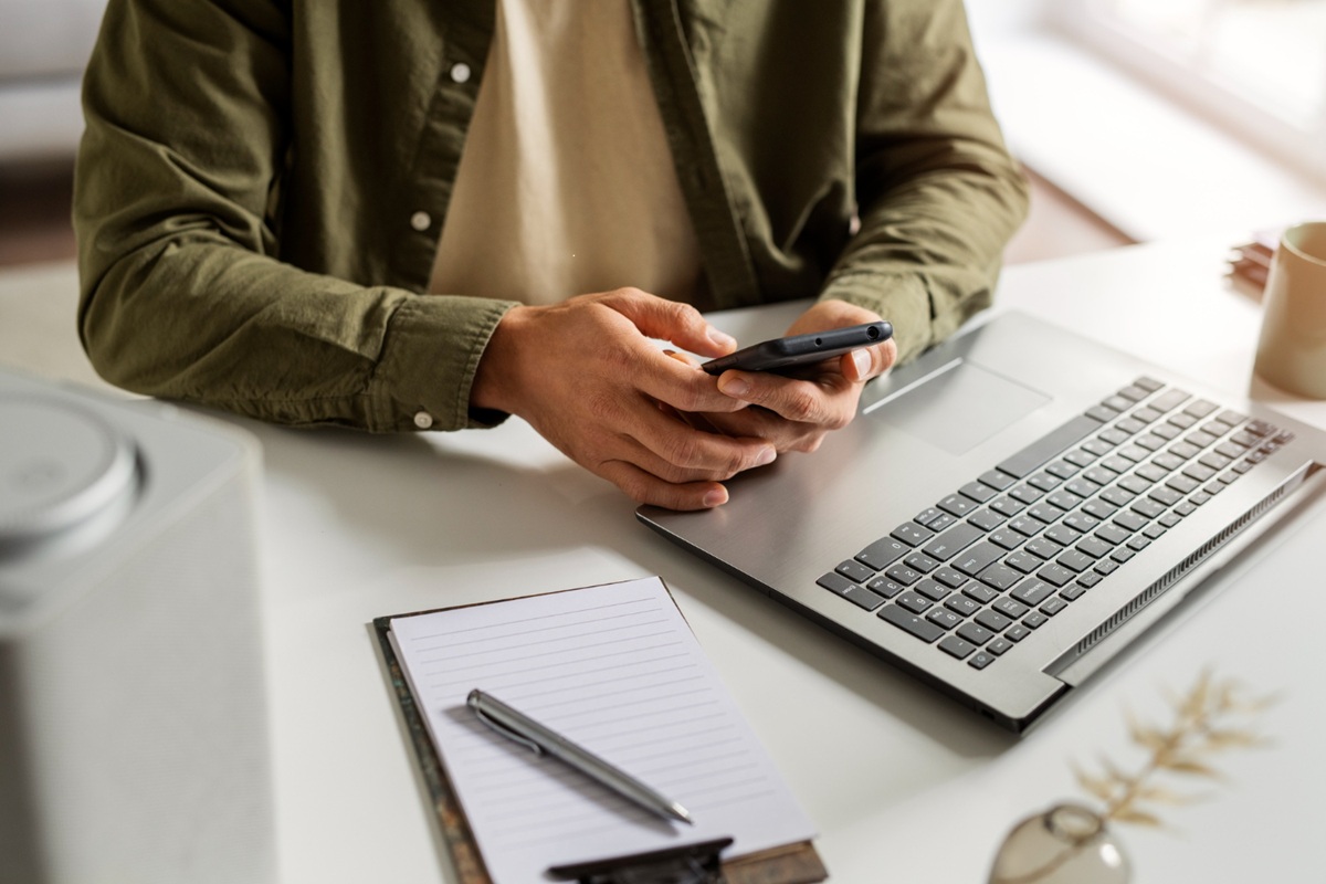 Homem usando celular sentado à mesa com laptop e bloco de notas ao lado, representando consultoria e mentoria.