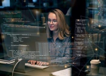 Mulher programadora trabalhando em computador, com código na tela e ambiente de escritório ao fundo.