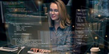 Mulher programadora trabalhando em computador, com código na tela e ambiente de escritório ao fundo.