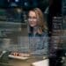 Mulher programadora trabalhando em computador, com código na tela e ambiente de escritório ao fundo.