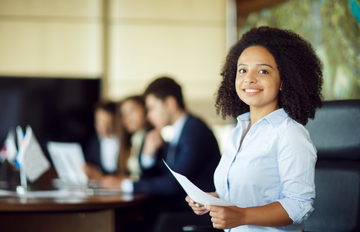 Jovem mulher sorridente em reunião de trabalho, com outras pessoas ao fundo desfocadas.