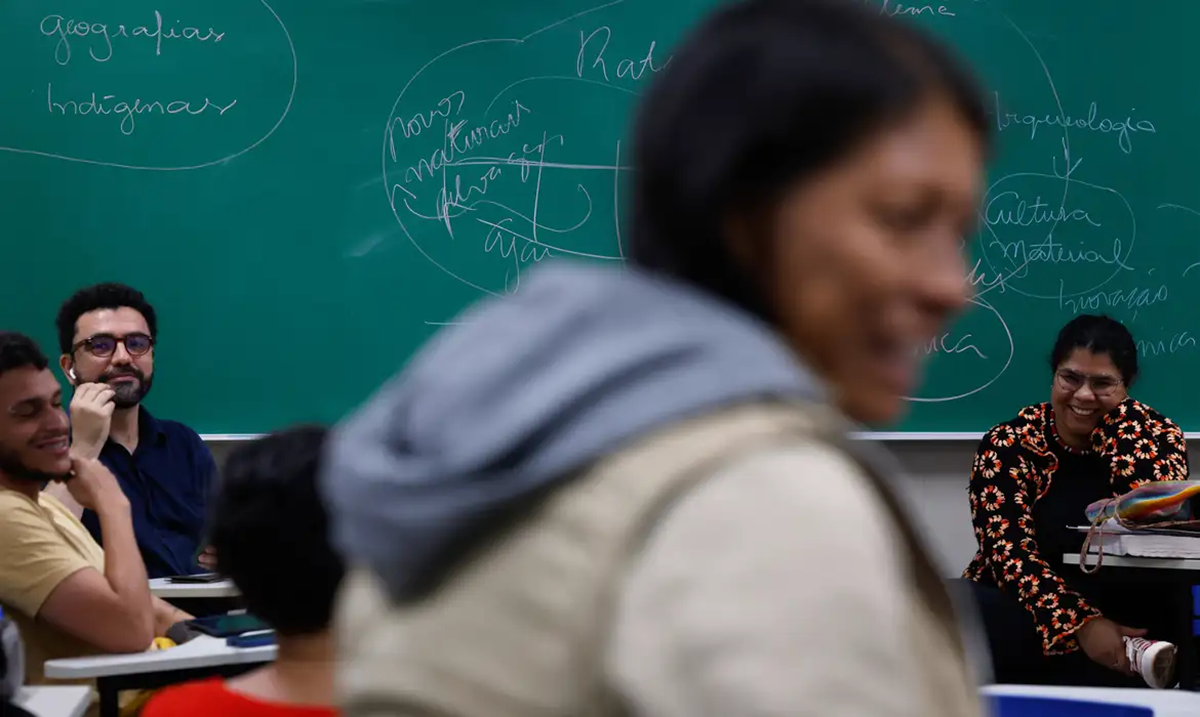 Sala de aula com alunos da EJA participando de discussão, com um quadro verde ao fundo.
