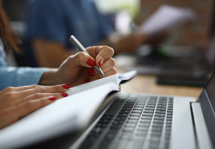 Mãos femininas escrevendo em um caderno sobre teclado de notebook, em ambiente de estudo ou trabalho com outras pessoas ao fundo.
