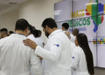Grupo de médicos em jalecos do SUS conversando em frente a um painel com mapa do Brasil e texto “Mais Médicos para o Brasil”