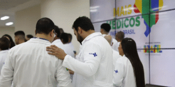 Grupo de médicos em jalecos do SUS conversando em frente a um painel com mapa do Brasil e texto “Mais Médicos para o Brasil”