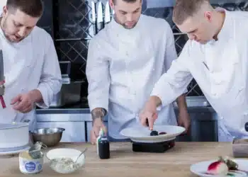 Três chefs trabalhando juntos em cozinha, com foco em pratos sofisticados.