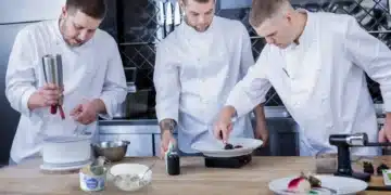 Três chefs trabalhando juntos em cozinha, com foco em pratos sofisticados.