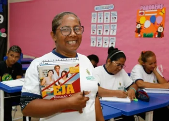 Aluna sorrindo enquanto segura material do EJA, com outros estudantes ao fundo."