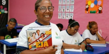 Aluna sorrindo enquanto segura material do EJA, com outros estudantes ao fundo."