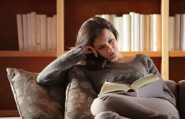 Mulher relaxada lendo um livro sentada no sofá com estantes de livros ao fundo