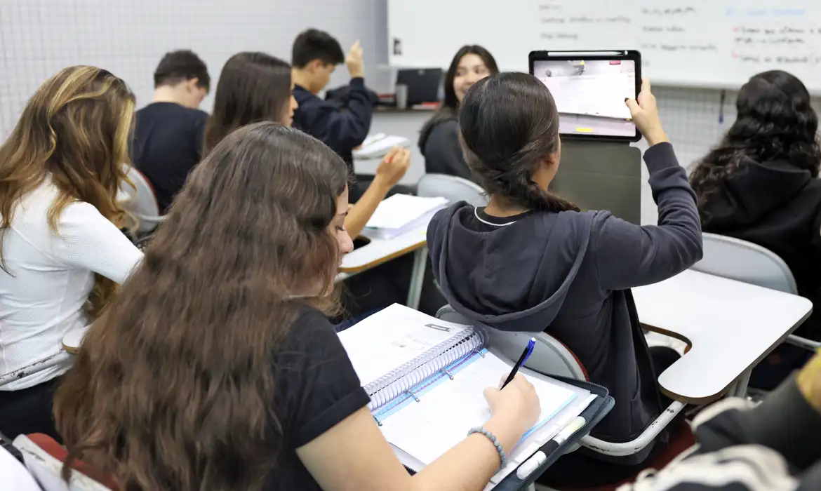 Alunos em sala de aula, alguns usando tablet e outros escrevendo em cadernos