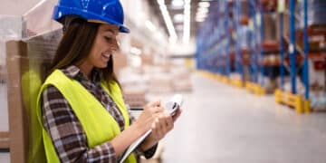 Mulher usando capacete e colete refletivo, escrevendo anotações em um ambiente de trabalho industrial.