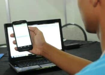 Jovem de camiseta azul segura smartphone na frente de notebook sobre mesa preta, em sala iluminada