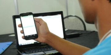 Jovem de camiseta azul segura smartphone na frente de notebook sobre mesa preta, em sala iluminada