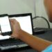 Jovem de camiseta azul segura smartphone na frente de notebook sobre mesa preta, em sala iluminada