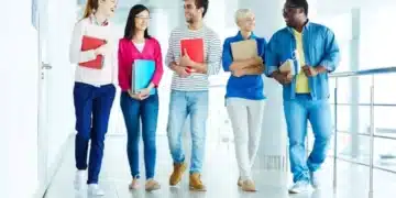 Grupo de estudantes caminhando pelo corredor de uma escola, sorrindo e segurando materiais de estudo
