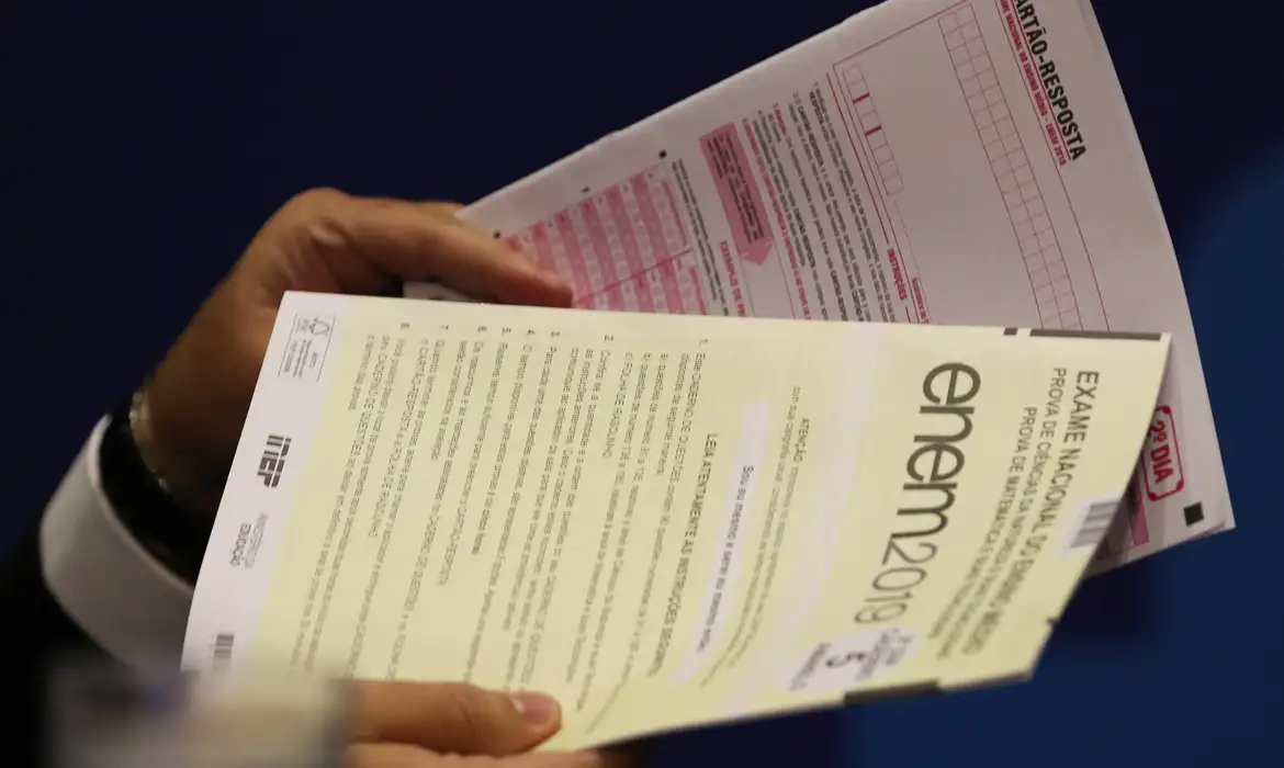 Pessoa segurando as provas do ENEM 2019, com destaque para o cartão de respostas e as instruções do exame.