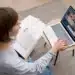 Mulher estudando online com videoaula no notebook e livro aberto ao lado