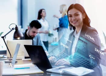 Imagem de um grupo de profissionais em um escritório moderno, com destaque para uma mulher sorridente em frente a um computador, envolvida com programação.