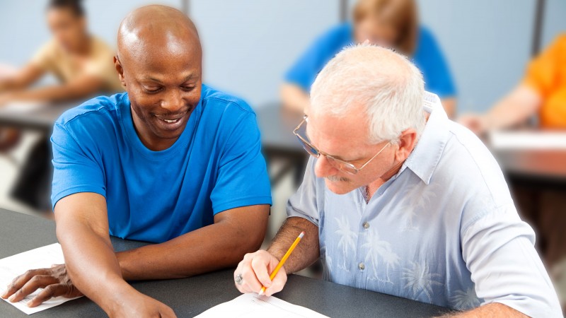 Estudar aos 50. Dois adultos estudando juntos em uma sala de aula, com um mais novo orientando o mais velho.