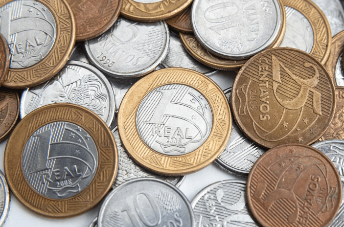 Moedas brasileiras de diversas denominações espalhadas, incluindo peças de R$1,00, 25 e 10 centavos