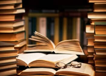 Foto detalhada de livros empilhados, com páginas abertas e óculos sobre um livro, simbolizando o estudo e a leitura.