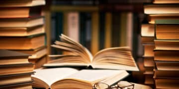 Foto detalhada de livros empilhados, com páginas abertas e óculos sobre um livro, simbolizando o estudo e a leitura.