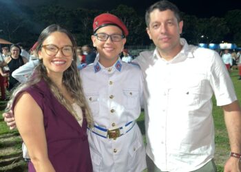 Lucca em uniforme, com seus pais, em evento ao ar livre à noite.