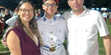 Lucca em uniforme, com seus pais, em evento ao ar livre à noite.