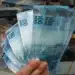Cédulas de 100 reais sendo seguradas por uma mão, com notas empilhadas ao fundo, representando riqueza e economia no Brasil.
