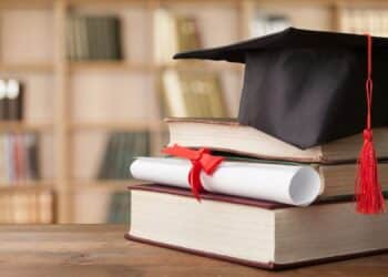 Imagem de um diploma enrolado com fita vermelha e um capelo de formatura, sobre uma pilha de livros